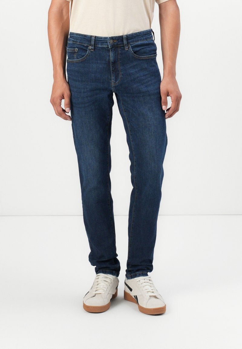 Vaqueros de mezclilla azul oscuro con un corte ajustado, que cuentan con cinco bolsillos y un lavado sutil, combinados con zapatillas blancas.