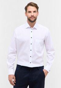 Weißes Langarmhemd mit spitzem Kragen, schwarzen Knöpfen und einer Brusttasche, getragen mit dunklen Hosen, glatte Stofftextur.
