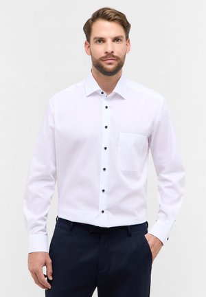 Witte lange mouwen shirt met een puntige kraag, zwarte knopen en een enkele borstzak, gedragen met donkere broek, gladde stofstructuur.