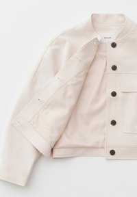 Lichtbeige jas met zwarte knopen, één borstzak en een ronde kraag, gedeeltelijk open getoond met zichtbare voering aan de binnenkant.