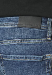 Jeans de mezclilla azul con una superficie texturizada, que presentan una etiqueta de cinturón de cuero negro marcada "ANTONY MORATO" y detalles de costura amarilla.