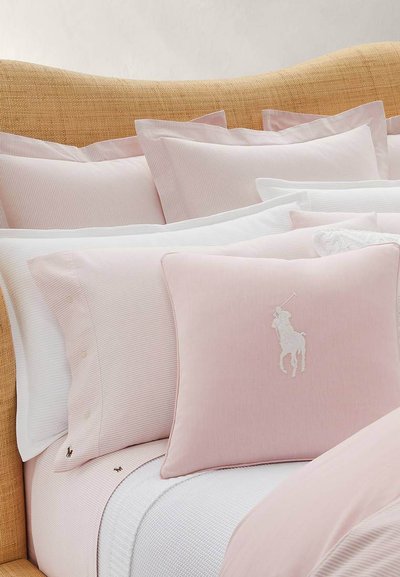 Απαλά ροζ και λευκά στρωμένα σεντόνια με μαξιλάρια που ταιριάζουν, συμπεριλαμβανομένου ενός διακοσμητικού μαξιλαριού με κεντημένο λογότυπο παίκτη πόλο.