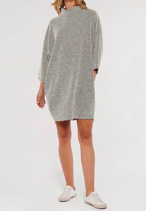 Femme portant une robe en maille gris clair ample et texturée avec des manches trois-quarts et des baskets blanches, debout sur un fond uni.