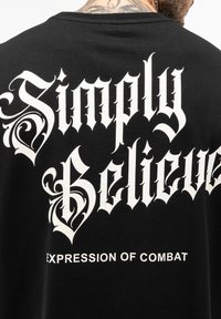 Črna majica s velikim belim gotičnim besedilom "Simply Believe" z dekorativnimi elementi in manjšim besedilom "EXPRESSION OF COMBAT" spodaj.