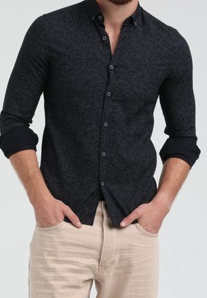 Man wearing a fitted black patterned button-up shirt with rolled sleeves and beige pants, hands in pockets, against a plain background.