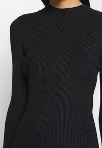 Pull noir côtelé à manches longues avec col montant porté par une personne aux longs cheveux foncés sur un fond uni.