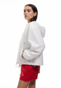 Witte capuchon windbreakerjack met elastische manchetten en verstelbare trekkoorden. Gecombineerd met rode shorts met zijzakken en borduursel.