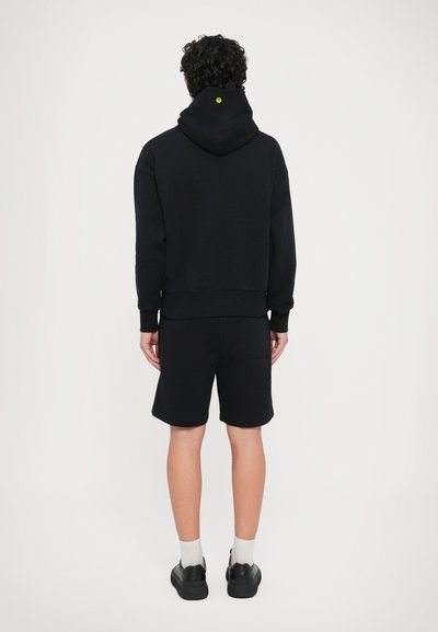 Melnā kapuce ar elastīgām aprocēm un mazu dzeltenu logotipu uz muguras, kopā ar saskaņotām melnām šortiem un melniem apaviem.