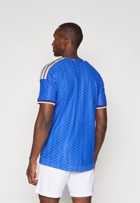 Plava atletska majica s teksturiranim uzorkom lista, kratkim rukavima i zlatnim detaljima na ramenima. Nosena preko bijelih šortsa.