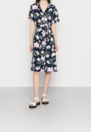 Blumenkleid in Marineblau, mit rosa und weißen Blumen. Lockere Passform mit kurzen Ärmeln, Wickeldesign und einem gebundenen Taillengürtel. Weiße Sandalen.