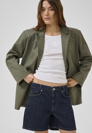 Nő, aki egy túlméretezett olívazöld blézert, fehér bordázott atlétafelsőt és sötétkék farmer rövidnadrágot visel, egyszínű háttér előtt.