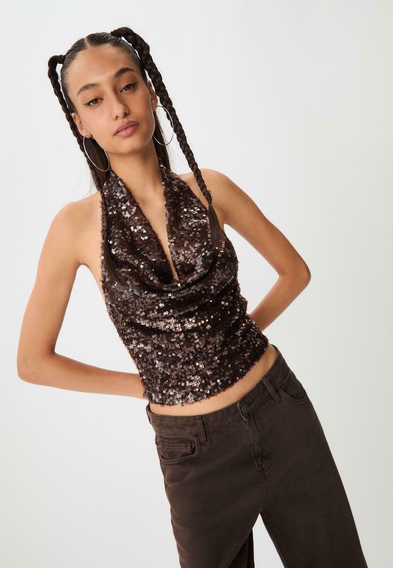 Hnedý halter top pokrytý flitrami s zloženým výstrihom, spárovaný s tmavohnedými nohavicami vysokej pásovky. Modelka pózuje s rukami umiestnenými popri tele.