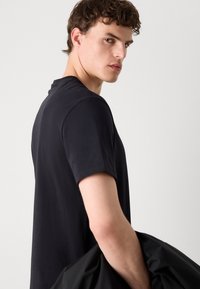 Ung mann med krøllete brunt hår iført en svart kortermet skjorte, holder en svart jakke, ser over skulderen.