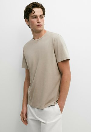 Junger Mann mit kurzen dunklen Haaren, der ein beiges Kurzarmhemd und weiße Hosen trägt, steht mit einer Hand in der Tasche und schaut seitlich.