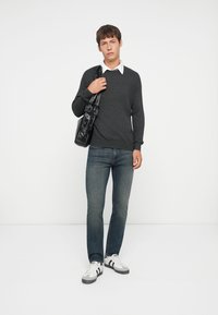 Grå ribbet genser, hvit skjorte under, blå jeans, svart crossover veske, hvite sneakers med sorte striper, står mot en ensfarget bakgrunn.