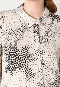 Camicia beige con motivo a pois neri, design con colletto, chiusura frontale con bottoni e tessuto leggero. Texture liscia, stile casual.