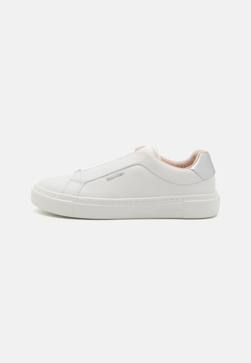 Balti slip-on sportbačiai su lygia odos viršūne, apvalia nosimi ir smėlio spalvos pamušalu. Sidabrinė detalė kulne, plokščias guminis padas, minimalūs siuvimo elementai.