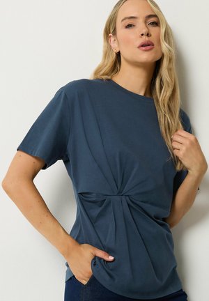 Donna con capelli biondi lunghi che indossa una maglietta blu scuro a maniche corte con dettaglio a torsione frontale, in piedi davanti a uno sfondo chiaro e uniforme.