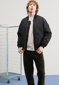 Giovane con capelli ricci che indossa una giacca bomber nera sopra una maglietta bianca e pantaloni scuri, in piedi accanto a un carrello di stoccaggio in filo metallico.
