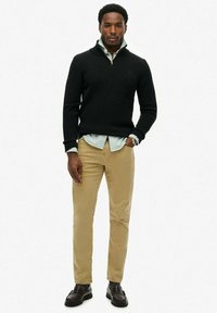 Czarny sweter z dzianiny z zamkiem przy szyi, zestawiony z jasnozieloną koszulą w paski i beżowymi spodniami. Czarne buty wizytowe dopełniają całość.