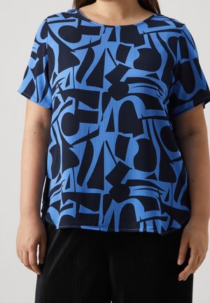 Kobieta w krótkim niebieskim T-shircie z abstrakcyjnymi czarnymi geometrycznymi kształtami, w połączeniu z czarnymi spodniami o fakturze.