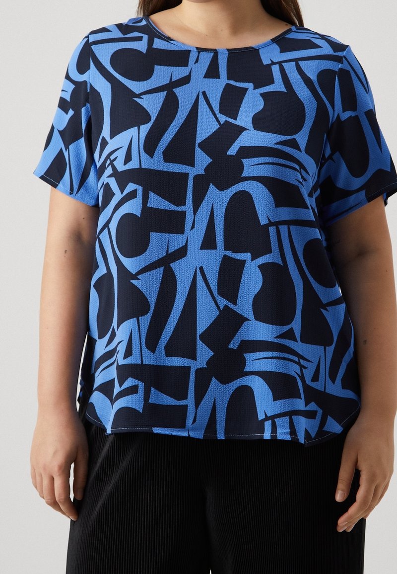 Femme portant une chemise bleu à manches courtes avec des formes géométriques abstraites noires, accompagnée d'un pantalon noir texturé.