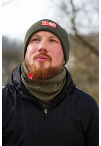 Olive green knit beanie with a red patch, matching ribbed neck gaiter, worn with a black zip-up jacket. Textured fabric, winter wear.