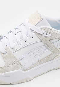 Weiße Sneaker mit Leder- und grauen Wildlederpaneelen, runden Schnürsenkeln und einer strukturierten Oberseite. Akzente sind ein sichtbares Stichmuster und eine graue Sohle.