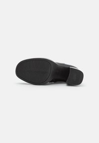 Semelle de chaussure en cuir noir avec un motif de grip texturé et un bout arrondi. Présente un petit talon carré et une légère courbure vers le talon.
