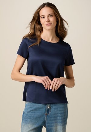 Kvinde med løst brunt hår iført en marineblå kortærmet skjorte og lyseblå jeans, stående mod en ensfarvet lys baggrund.