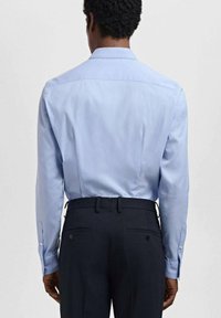 Lichtblauw lang shirt met een ruggegot en knopenmanchetten, gecombineerd met donkere broeken met twee achterzakken en knopen.