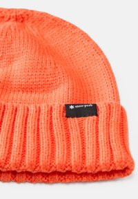 Knit orange beanie with a ribbed cuff, featuring a black label with white text. Soft texture, warm design suitable for colder weather.
