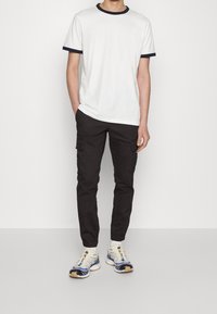 Biała koszulka z okrągłym dekoltem i granatowym obszyciem, w połączeniu z czarnymi spodniami cargo z bocznymi kieszeniami oraz beżowo-niebieskimi butami sportowymi o teksturowanym wzorze.