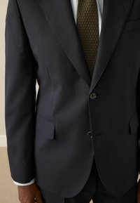 Dunkelblauer Anzugjacke aus feinem Stoff, mit abgesetztem Revers, zwei Knöpfen und einer einzelnen Brusttasche; darunter eine strukturierte bronzefarbene Krawatte.