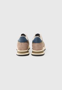 Ζευγάρι μπεζ, καφέ και μπλε αθλητικά παπούτσια απεικονιζόμενα από πίσω με μαύρες και κρεμ σόλες σε ανοιχτό γκρι φόντο.