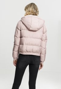 Kvinna som bär en ljusrosa vadderad jacka med huva och svarta tajta jeans, stående med ryggen mot en enfärgad ljus bakgrund.