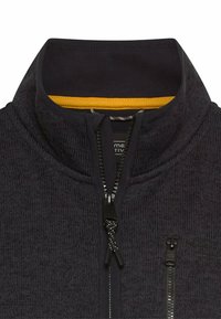 Dunkel gestrickte Fleecejacke mit hohem Kragen, ausgestattet mit einem gelben Akzent innen am Kragen und einer aufgesetzten Brusttasche mit Reißverschluss. Schwarze Reißverschlussbeschläge.