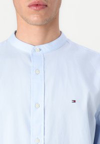 Mand iført lyseblå skjorte med mandarinkrave, hvide knapper og lille Tommy Hilfiger-logo på venstre bryst.