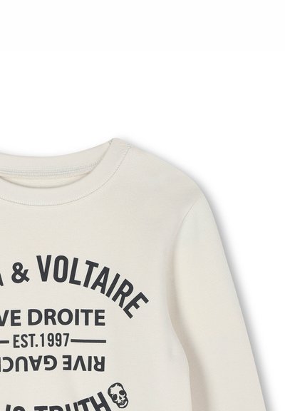 Lichtbeige trui met een ronde hals, voorzien van vetgedrukte zwarte tekst en een schedelafbeelding. De stof lijkt zacht met een lichte textuur.