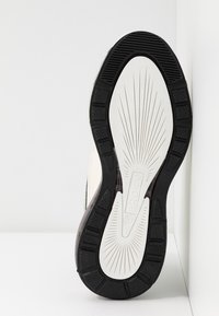 Semelle d'une basket noir et blanc avec une surface texturée et un motif en rayons de soleil, affichant le nom de la marque "BRONX" sur la partie blanche.
