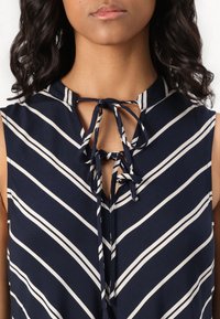 Blusa sem mangas azul-marinho com riscas brancas diagonais, apresentando um decote em forma de buraco de chave e um fecho amarrado à frente. Textura de tecido suave.