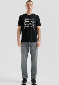 Schwarzes Baumwoll-T-Shirt mit metallischem Aufdruck des Wortes "METALLICA" in mehrfacher Ausführung. Getragen mit grauen Jeans und schwarzen Sneakers.
