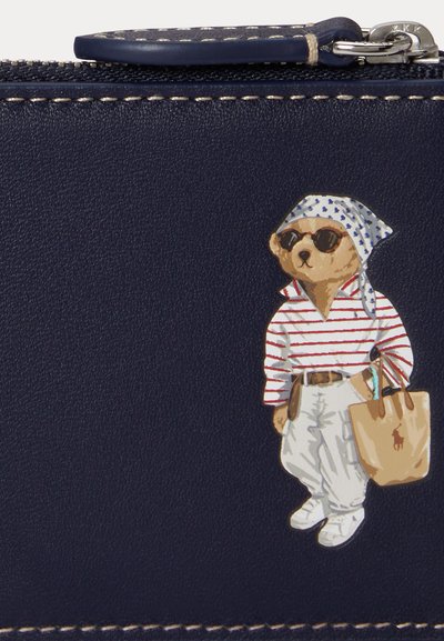 Un ours portant des lunettes de soleil, une chemise rayée, un pantalon blanc, un foulard sur la tête et qui porte un sac cabas beige sur une surface texturée bleu marine avec un détail de fermeture éclair.