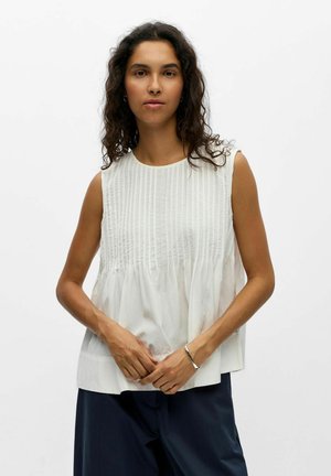 Femme aux longs cheveux bouclés portant un haut blanc plissé sans manches et un pantalon foncé, debout les mains jointes devant elle.