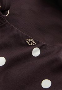 Brauner Stoff mit großen weißen Punkten und einem strukturierten schwarzen Netzpanel. Enthält ein kleines, rundes Metall-Logo-Detail.