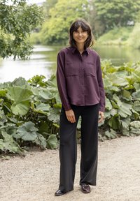 Camicia button-up color marrone scuro con tasche sul petto, abbinata a pantaloni larghi neri e mocassini neri lucidi. Foliage verde e acqua sullo sfondo.