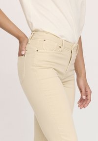 Jean taille haute beige au design ajusté, avec des poches avant, des coutures décoratives et des accents en métal.