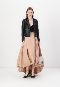 Giacca biker in pelle nera sopra una maglia nera, abbinata a una gonna beiger chiara con un taglio asimmetrico e scarpe con tacco alto nere. Borsa bianca semplice.