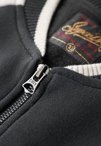 Sweatshirt noir à fermeture éclair avec un col côtelé contrastant, doté d'une fermeture éclair argentée gravée de "DRY" et d'une étiquette à l'intérieur.