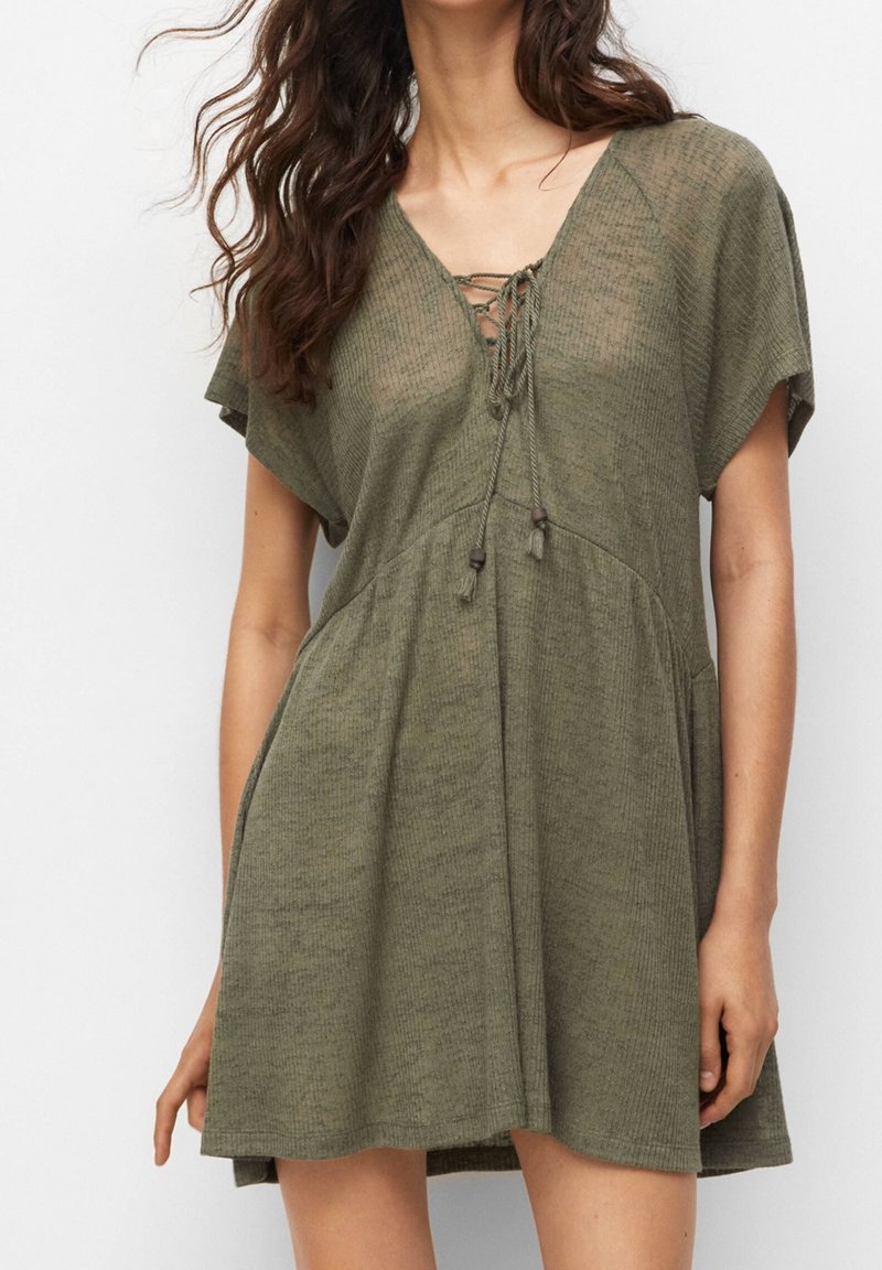 Femme portant une robe ample kaki à manches courtes avec un col en V lacé et des poches, debout devant un fond clair uni.